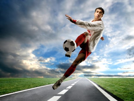 Man Wearing White Long Sleeved Shirt And Red Shorts Kicking Soccer Ball On The Road Below White Clouds And Blue Sky During Daytime
