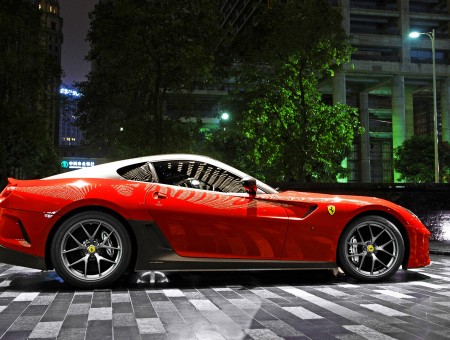 Red Ferrari Sports Car Parked Near Green Trees And Buildings At Night