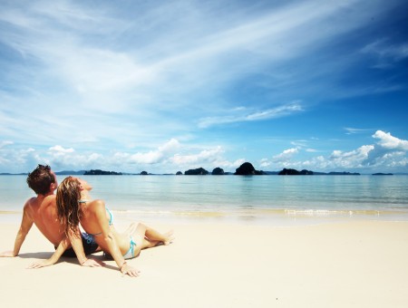 Man And Woman Sitting On Brown Sand Near Body Of Water Under Sunny Blue Cloudy Sky