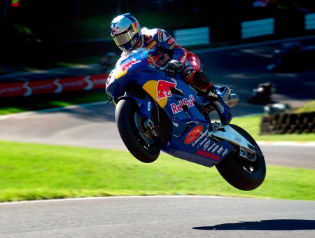 Man Wearing Blue Red And Black Jacket Riding On Blue Red And Yellow Sports Bike Jumps Over Gray Track During Daytime