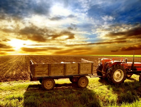 Red And Black Lawn Tractor Carrying Brown Frame 4 Wheel Utility Trailer In Front Of Fields Under Cloudy Orange And Blue Sky During Sunset